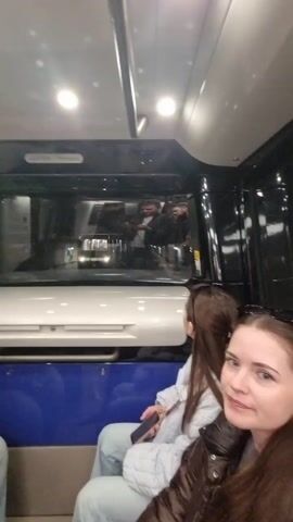 Two young women sit inside a metro train. The woman closest to the camera looks directly at the camera. The other woman looks out the window into the driver's cabin. The window shows the reflection of the driver. There is a white panel below the window, and a blue seat back in front of the women. The ceiling is silver and dotted with lights. The train car has black walls.
