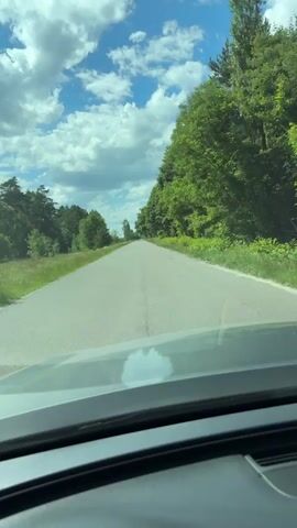 The video shows a point-of-view shot from inside a car traveling down a paved road. The road is surrounded by green trees on the right side and a grassy field on the left. The sky is blue with scattered white clouds. The dashboard of the car is visible in the foreground. The video appears to be a short clip, possibly a few seconds long.