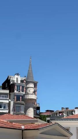 The video shows a minaret, a tall, slender tower attached to a mosque, with modern buildings in the background. The minaret is made of stone and has a conical roof topped with a golden finial. The buildings behind it are a mix of old and new, with some featuring balconies and large windows. The sky is a clear, bright blue, suggesting a sunny day. The terracotta-tiled roofs of the foreground buildings contrast with the smooth, white facades of the modern structures. There are no people visible. The scene appears to be set in Beşiktaş, Turkey, a district known for its historical sites and vibrant urban life. The time of day is likely midday, given the strong sunlight. The overall impression is one of a serene urban landscape, where historical architecture is integrated with contemporary development.