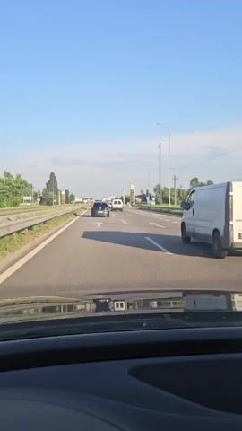 A first-person perspective shot from a vehicle driving on a highway. The road is paved and appears to be a multi-lane highway. There are other vehicles on the road, including cars and vans. The environment appears to be rural or suburban, with trees and vegetation visible on the sides of the road. The sky is clear and blue, suggesting fair weather. The time of day appears to be morning or late afternoon, given the angle of the sun and the lighting conditions.