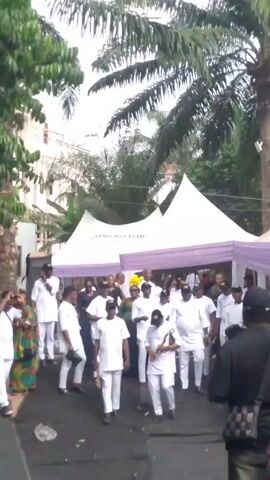 A crowd of people mostly dressed in white formal attire is walking down a street, some carrying small objects, toward several white tents with purple trim.  A palm tree towers to the right of the tents, and a building can be seen in the background. The person filming is wearing a colorful shirt and a black baseball cap with a white Nike logo. The day appears overcast.