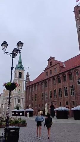 The video shows a historic European city square with cobblestone pavement. In the foreground, two young women walk away from the camera, dressed in casual summer clothing. Behind them, a tall, red brick building with a clock tower dominates the scene. The clock tower has a large clock face and a Polish flag at its peak. In front of the tower, there are market stalls with striped awnings. To the left of the main building, a green spire of another church can be seen. To the right of the clock tower, a bronze statue stands on a pedestal, next to a fountain. In the background, a row of colorful, multi-story buildings line a street, some with illuminated shop windows and outdoor seating areas. The sky is overcast, suggesting a cloudy or late-day setting. There are a few other people scattered around the square, some sitting and others walking. A street lamp with multiple fixtures is visible on the left side of the frame. The overall atmosphere is calm and somewhat subdued, likely due to the weather or time of day.