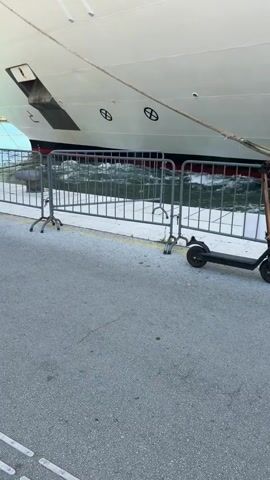 The video shows a person on a scooter parked next to a large white ship docked at a pier. The scooter is dark gray and has a black storage box on the back. The person is wearing a white t-shirt, a white baseball cap, and olive green cargo shorts. They are facing away from the camera and looking towards the ship. Metal barriers are set up between the scooter and the ship. The water around the ship is choppy.