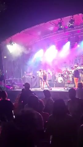 This is a first-person perspective video of a live music performance at night. The stage is lit with vibrant pink and blue lights, with smoke effects creating a hazy atmosphere. A band is performing on stage, with several musicians playing instruments like guitars, drums, and possibly a saxophone. A female singer is in the center of the stage, wearing a dark dress. A banner with text that appears to read "VIROWE NATION FE" is visible behind the band. In the foreground, the silhouettes of a crowd of people are visible, facing the stage and watching the performance. The overall atmosphere is that of an outdoor concert or festival.