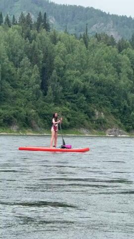 A young woman is stand-up paddleboarding on a wide, calm river. She is wearing a life vest over a swimsuit and holding a paddle. A pink bag is secured to the front of her bright orange paddleboard. The paddleboard is positioned in the center of the frame, with the woman facing left.

The river is surrounded by a lush, green, forested mountainside. The trees are dense and varied, with a mix of deciduous and coniferous species. The sky is overcast, suggesting a cloudy or possibly rainy day. The water appears slightly choppy with ripples, indicating a gentle current or light wind.

In the distance, on the right side of the frame, another person can be seen on the water, possibly also paddleboarding or in a small boat, though they are too far away to be clearly identified. The overall scene is serene and natural, evoking a sense of outdoor recreation in a picturesque, remote location. The time of day is likely daytime due to the ambient light, but the overcast sky prevents any strong shadows. The location is identified as Ust-Mana, Russia. There is no visible text in the media file.