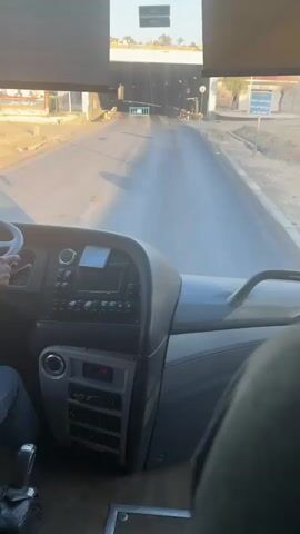 The video shows a first-person perspective from inside the front of a bus, looking forward through the windshield. The driver's hands are visible on the steering wheel on the left, and part of a passenger's shoulder or seat is on the right. The bus's dashboard and control panel are prominently displayed in the foreground.

Outside, the bus is on a paved road, approaching what appears to be an entrance to a tunnel or underpass made of concrete. The sun casts long shadows on the road, suggesting it's either morning or late afternoon. A blue barrier with Arabic text is positioned in the middle of the road ahead, and a red traffic cone is visible on the right side. A small green light is also visible on the right, possibly indicating entry clearance. The overall scene suggests the bus is navigating an entry point in Abu Sial, Egypt.