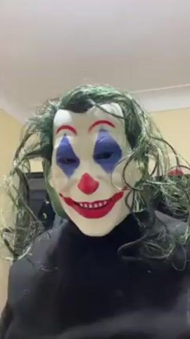 A person wearing a Joker mask and a black turtleneck stands indoors against a plain beige wall and waves. The mask is pale with red lips, a red clown nose, blue diamonds around the eyes, and curly green hair. The room is dimly lit by overhead lights.