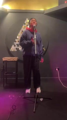 A young person stands singing into a microphone on a dimly lit stage. They are wearing a blue hoodie over a red hooded shirt and black pants with white sneakers. The background features a black wall with a circular decoration made of reflective discs arranged in a triangle shape. To the left, a stool with a textured seat is visible. Pink and purple lights illuminate the stage. A black microphone stand with a microphone is positioned in front of the singer. A white power cord is plugged into a wall socket on the right side of the frame. The singer's right hand is resting on their chest, and their left arm is slightly extended to the side. The singer's head is tilted back as they sing.