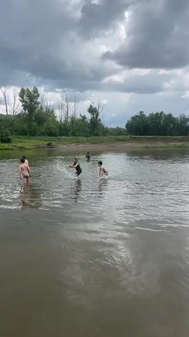 The video shows several children playing in a river. The water is murky and shallow enough for the children to stand. In the foreground, the water is rippling. In the background, there is a tree line with green foliage and some bare branches. The sky is cloudy and overcast. The children are spread out in the river, some of them interacting with each other. One child is in the center of the frame, wearing a black shirt and dark bottoms, and appears to be kicking their legs. Another child, to the right, is wearing light-colored swim trunks and is facing the child in the black shirt. To the left of the frame, there are two other children in swim trunks. The overall impression is one of casual, outdoor recreation.