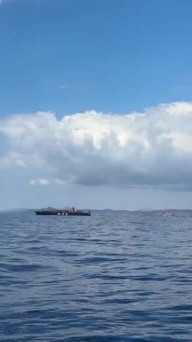 The video captures a serene daytime scene of the open sea under a partly cloudy sky. A large container ship, clearly marked with "MSC" on its side, sails from left to right across the mid-ground. The ship is heavily loaded with numerous shipping containers stacked high on its deck.

The sky is a bright blue, partially covered by expansive white and grey cumulus clouds that stretch horizontally across the upper portion of the frame. The water is calm, a deep blue, with gentle ripples and reflections visible on its surface. In the far distance, a low, indistinct grey-brown landmass, likely a coastline or islands, can be seen beneath the cloud cover.

The setting is an expansive marine environment, possibly a shipping lane or coastal waters. No people are visible. The video evokes a sense of peacefulness and the vastness of the ocean, with the large vessel making its journey. The ambient sound appears to be that of water, possibly waves or a boat moving. The provided context indicates the location is Koundouros, Greece.
