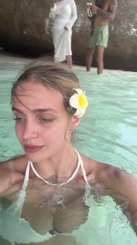 A young woman is taking a selfie video while standing in clear, light-green water. She is wearing a white bikini top and a necklace made of white beads. A white flower is tucked behind her ear. Her hair is wet and pulled back, and she smiles and winks at the camera. In the background, there are other people standing on a sandy beach and large rocks. One person is wearing a long white dress. The scene appears to be a tropical beach setting.