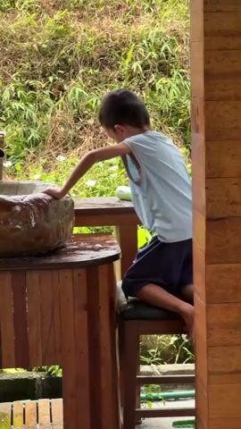 A young boy is captured from behind, engaged in an activity at a rustic outdoor washing station. He wears a light blue sleeveless top and dark shorts, with his short dark hair visible. The boy is leaning forward, with his left arm extended into a large, irregular-shaped basin that appears to be made of stone or carved wood, filled with water. This basin is mounted on a wooden counter. A wooden stool is positioned to the left of the counter, which the boy is standing next to or on to reach the basin.

In the background, there is a dense expanse of lush green foliage, including bushes and ferns, indicating a natural, outdoor setting. A small green bottle, possibly containing soap, rests on the counter behind the basin. On the right side of the frame, a wooden pillar or wall is visible, suggesting part of a structure like a house or a shelter. The overall impression is one of a simple, natural environment. The light appears to be natural daylight, and the greenery suggests a damp or humid climate. No specific text is visible in the frame. The scene is consistent with an outdoor area in Du Già Commune, Vietnam.