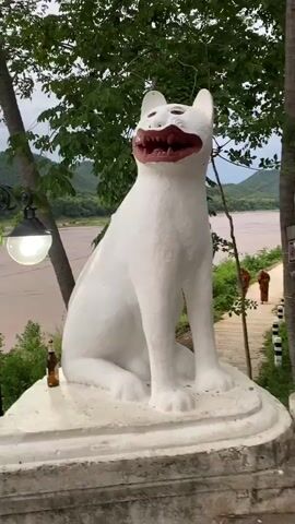 A white cat statue with its mouth open and teeth showing sits in the foreground. Behind the statue, two Buddhist monks in orange robes walk down a path next to a river. The river is wide and muddy, with green hills in the background. Trees and foliage surround the scene.