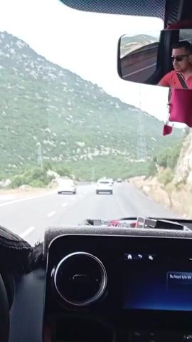 The video shows a view from inside a vehicle traveling on a highway. The driver is visible in the rearview mirror, wearing a red shirt and sunglasses. The highway passes through a mountainous, green landscape. There are other vehicles on the road ahead, and power lines are visible along the side of the road. The dashboard of the vehicle is partially visible in the foreground.
