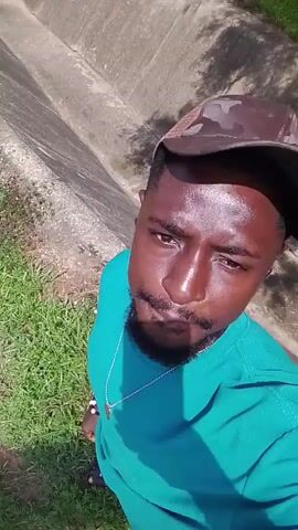 An adult male with dark skin and a beard, wearing a teal long-sleeved shirt, a camouflage baseball cap, a silver chain with a pendant, and a black earbud in his right ear, is filmed outdoors. He slowly turns his head to look directly at the camera, maintaining eye contact, and briefly opens his mouth at the end of the clip. The background consists of green grass and a concrete structure with dirt. The scene is brightly illuminated by sunlight.