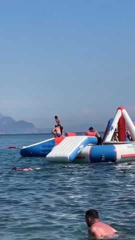 The video shows a sunny day at a beach with several inflatable water toys in the ocean. In the foreground, two people are in the water. One is a man, partially submerged, facing away from the camera. Another is a child wearing goggles and a pink swimsuit, swimming towards the camera. In the background, a large inflatable water park structure is visible. One person is on top of the structure, and another is in the process of sliding down a blue and white ramp, splashing into the water. Several other people are scattered on and around the inflatable structures. The water is clear blue, and in the distance, mountains can be seen under a clear sky.