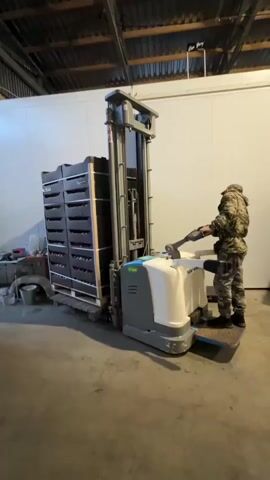 A man wearing camouflage clothing and a cap operates a powered pallet stacker. The stacker is lifting a pallet stacked with dark crates. The man is standing on the operator platform of the stacker, holding the controls. The scene appears to be inside a warehouse or storage facility, with large white insulated walls and a corrugated metal ceiling. Other pallets of crates are visible in the background. The lighting is artificial and casts shadows on the concrete floor.