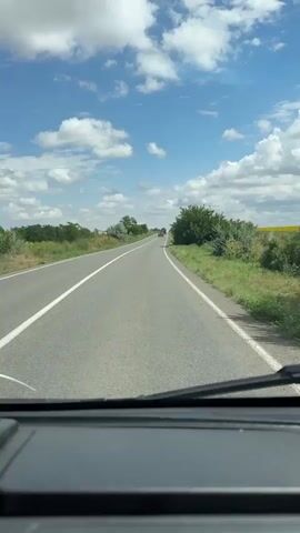 This is a first-person perspective video of a person driving a vehicle on a road. The view is from inside the vehicle, looking out through the windshield and side mirror. The road is paved with a white line on the left side and a white dashed line on the right. The surrounding environment is rural, with green grass and trees on both sides of the road. There are a few other vehicles visible in the distance. The sky is blue with scattered white clouds. The video transitions to a close-up of the side mirror, showing the vehicle's reflection and the road behind it. The vehicle has a white body with red stripes.