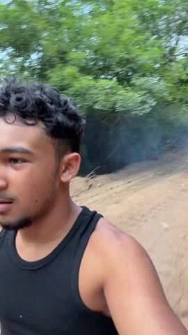A young man wearing a black tank top and olive green shorts is riding an off-road vehicle. The vehicle appears to be a dirt bike or ATV, judging by the handlebars visible in the foreground and the rider's posture. He is moving at a fast pace on a dirt path, with trees and foliage on either side of the path. There is a plume of blue smoke trailing behind the vehicle, suggesting it may be a smoke bomb or a malfunction. The rider appears to be focused on the path ahead, with a determined expression on his face. The environment is outdoors, likely a rural or wilderness area, with dry dirt and vegetation visible. The lighting suggests it is daytime, with natural light filtering through the trees.