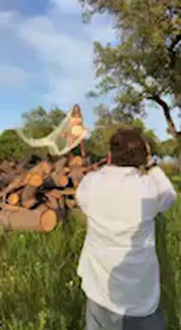 In a sunlit outdoor setting at El Chaparrito, Spain, a model and a photographer are engaged in a photoshoot. The scene features a large pile of cut logs and wood, with green grass in the foreground and various trees, including a prominent one on the right, forming the background against a blue sky with scattered clouds.

A woman, dressed in a light-colored, possibly white or cream-colored, two-piece outfit that appears to be a bikini or similar attire, stands atop the pile of logs. She is holding and artfully posing with a large piece of flowing white fabric or shawl. She extends her arms, allowing the fabric to billow around her, creating dynamic shapes. Her attention is directed towards the photographer.

A man, seen from behind and wearing a white shirt, is positioned in the foreground, facing the woman. He holds what appears to be a camera, actively taking photos of the model. The video captures a brief moment of this creative process, showcasing the model adjusting her pose with the fabric and the photographer capturing the shots. The overall atmosphere suggests a professional, outdoor fashion or art photoshoot.