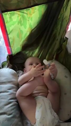 A baby is lying on its back in a playpen. The baby is wearing a diaper and has its legs raised in the air. The baby is holding a toy in its hands and appears to be playing with it. The playpen has a camouflage-patterned fabric lining. A pillow is visible next to the baby. The video is shot from a close-up perspective.