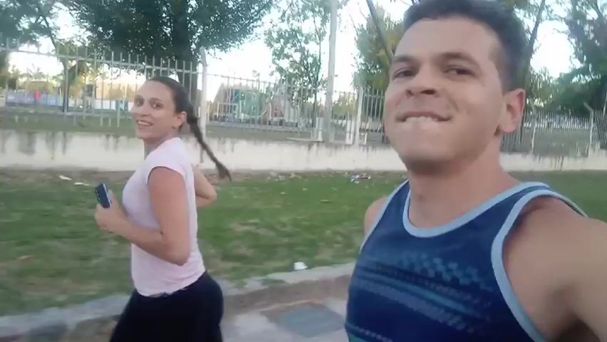 A man and a woman are jogging together. The man is holding the camera, so it’s a first-person perspective. He is wearing a dark blue tank top. The woman is wearing a pale pink t-shirt and dark leggings and has her hair tied back. The woman looks at her phone. They are jogging on a paved path with grass and a metal fence behind them. Trees are visible in the background. The man looks forward and smiles and talks.