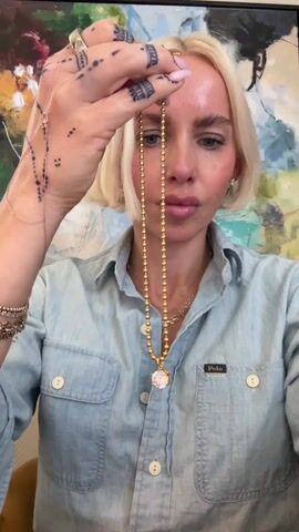 A blonde woman is centered in a frame, holding up a gold chain necklace with a white pendant. She is wearing a light-wash denim shirt with a Polo logo. She has tattoos on her hands. She is indoors, with a large colorful abstract painting behind her. She appears to be fastening the necklace. She is also wearing another gold necklace.