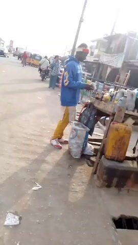 A man in a blue and yellow sweatshirt, yellow pants and a red-and-white hat stands behind a wooden table lined with containers of various substances, possibly liquids. He is looking down at the table, potentially arranging items. A woman with long, dark hair walks past him, carrying a black tote bag. The scene appears to be a busy street or market area with buildings and vendors nearby. There are also several sacks on the ground near the wooden table.