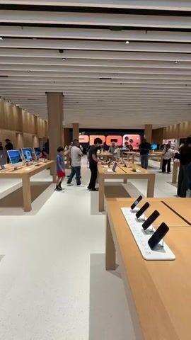 The video shows the interior of a modern retail store, likely an electronics store, with bright lighting and minimalist design. The floor is a light-colored speckled material, and the ceiling features a slatted design. Wooden display tables are arranged throughout the space, showcasing various electronic devices. In the foreground, a table displays several smartphones in charging stands. Further back, a group of people are gathered around display tables where laptops and other devices are exhibited. Several individuals are interacting with the products, and some appear to be employees assisting customers. In the background, a large digital display screen shows promotional imagery, possibly for a new product launch, with the text "iPhone 17 Pro" visible. The overall atmosphere suggests a busy retail environment.
