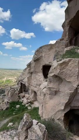 This video showcases a large, light-colored rock formation featuring numerous carved openings, appearing to be ancient cave dwellings or rooms. The rock face is rugged and uneven, with visible strata and natural erosion marks. In the background, a blue sky with scattered white clouds is visible, and to the left, a distant, arid valley with sparse vegetation stretches under the bright daylight.

As the camera pans downwards, more of the rock formation's base is revealed, showing large, weathered boulders and patches of green grass and small shrubs growing amidst the rocks. Several more carved entrances are visible at ground level, some partially obscured by the fallen rocks. The camera then zooms into one of these lower openings, providing a closer look at the dark interior of what appears to be a cave entrance or a room carved directly into the rock. The rugged texture of the rock surface around the entrance is prominent. No people or specific activities are depicted in the video. The scene evokes a sense of historical dwelling or natural wonder in a dry, open landscape, consistent with the provided location context of Acıgöl, Turkey, which is known for its unique geological formations and ancient settlements.