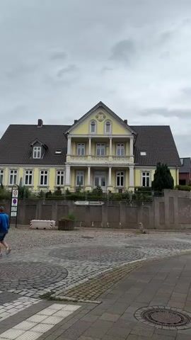 The media file shows a two-story yellow building with a dark roof and a prominent central balcony supported by columns. The building has numerous windows, and a triangular pediment sits above the balcony on the top floor, featuring decorative elements and what appear to be arched windows. The building is set behind a low, stone-like wall and a fence. In front of the building, there is a cobblestone street with a pedestrian crossing.

Visible objects include traffic signs, a concrete barrier, and planters with some greenery. The sky is overcast, suggesting a cloudy or possibly rainy day. The overall scene appears to be a street view in a town or city.

Text visible on a sign in front of the building includes "Evangelisches Krankenhaus" and a red cross symbol, indicating that this is likely the Evangelical Hospital. Another sign visible on the left appears to be a traffic sign.

No people are clearly visible in the frame. The activity depicted is primarily static, focusing on the architecture of the building and its immediate surroundings. The setting appears to be a public area, likely a street or square in front of the hospital. The mood is neutral, conveyed by the overcast weather and the absence of people. The location is identified as Bassum, Germany.