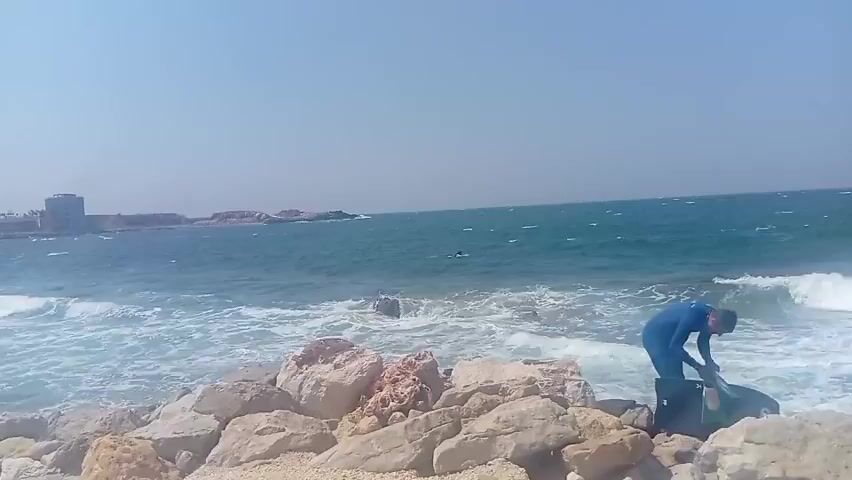 A person in a blue wetsuit walks along a rocky shoreline carrying a black bodyboard. The person is walking towards the left of the frame. Waves are crashing on the shore, and the sea is a deep blue. In the distance, there are a few other people in the water and a building on a rocky outcrop. The sky is clear and blue.