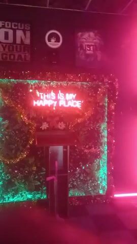 The video shows a room with a neon sign that reads "THIS IS MY HAPPY PLACE" surrounded by greenery and string lights. Above the sign is a poster with the words "FOCUS ON YOUR GOAL." To the left of the sign is a white refrigerator. A neon sign reading "RED BOX" can be seen above the refrigerator. The room appears dimly lit with red and green lighting.