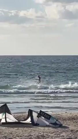 The video shows a person kitesurfing in the ocean. The person is dressed in a dark wetsuit and is holding onto a kite, which is not visible in the frame. The person is riding the waves and moving across the water. In the foreground, on the beach, there is a kite laid out on the sand. The sky is light blue with white clouds. The water is choppy.