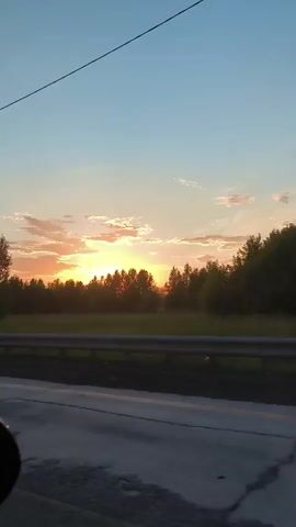 A sunset viewed from a car window shows the sun setting behind a line of trees, casting an orange glow on the clouds. The sky is a gradient of blue and orange. A highway guardrail is visible in the foreground, separating the road from a field. A utility pole is also visible on the right side of the frame.