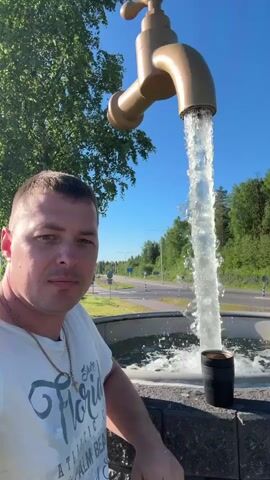 A man in a white t-shirt stands in front of a large faucet sculpture that is spraying a powerful stream of water into a fountain. The man is looking at the camera. In the background, there is a road with trees and a blue sky. The man has a gold chain around his neck and the t-shirt has "Florida" written on it in large letters. A black cup is visible in the fountain.