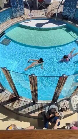 The video shows an elevated view of a pool area on what appears to be a cruise ship. In the foreground, two women are swimming in a small, round pool with clear blue water, bordered by a shallow, light green section. The pool is surrounded by a glass and metal railing. A man is sitting on a deck near the pool, looking down at his feet.

In the background, a larger rectangular pool is visible, with a red umbrella providing shade for a lifeguard sitting in a chair. The deck surrounding the pool has lounge chairs and several people relaxing. The upper deck of the cruise ship is visible, with various architectural features. Beyond the ship, there is a cityscape visible under a clear, sunny sky.