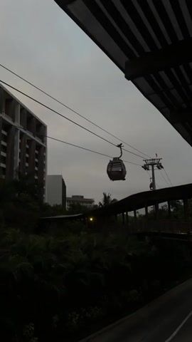 The video shows a cable car slowly moving through the air, viewed from underneath a wooden walkway structure with an open-slatted roof. The sky is overcast and the light is dim. In the background, there are buildings, trees, and more sections of the wooden walkway. As the camera moves forward along the walkway, people become visible walking on it. The view is slightly obscured by the support pillars of the walkway structure. The overall impression is a quiet, still scene in a slightly exotic location.