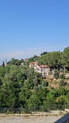 The video shows a wide shot of a hilly landscape with lush green trees and houses scattered on the slopes under a clear blue sky. In the foreground, there's a paved area with a fence. A white mosque minaret is visible in the distance on one of the hills. As the camera pans slightly to the left, a city appears in the far distance, nestled in a valley, with mountains forming the backdrop.