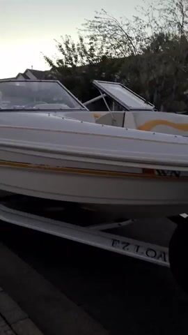 A white boat with yellow and black accents is parked on a trailer. The boat has a windshield and what appears to be a white Bimini top. The trailer is dark and appears to be made of metal. The boat is situated on a paved surface, possibly a driveway or street, with a sidewalk visible in the foreground. Behind the boat, there are houses with pitched roofs and trees. The sky is a pale blue with some wispy clouds, suggesting it might be dusk or dawn. The lighting is dim, casting soft shadows. There are no people visible in the video.
