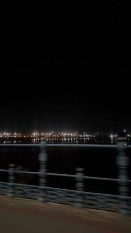 The video shows a night scene with a view of a waterfront cityscape. The camera appears to be moving, likely from a vehicle, as evidenced by the passing guardrail of a bridge or road. The night sky is entirely dark, contrasting with the numerous lights along the distant shoreline, which are reflected in the calm water. These lights likely originate from buildings, streetlights, or other urban fixtures. The guardrail is made of a dark metal and has a classic, ornate design.  The scene is mostly static except for the slight movement of the camera's viewpoint.
