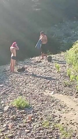 Two individuals are standing on a rocky riverbank under bright sunlight. The person on the left, appearing to be a female with pink hair, is wearing a dark swimsuit and holding a towel to her head. The person on the right, appearing to be a male, is shirtless and wearing dark shorts. He is holding a blue object, possibly clothing or a bag. In the background, there is dense foliage and a dark, possibly forested, hillside. Scattered on the riverbank are several dark objects, possibly bags or clothing, and some scattered debris, including a white plastic bottle. The river itself is visible in the lower portion of the frame, with calm water flowing over rocks.