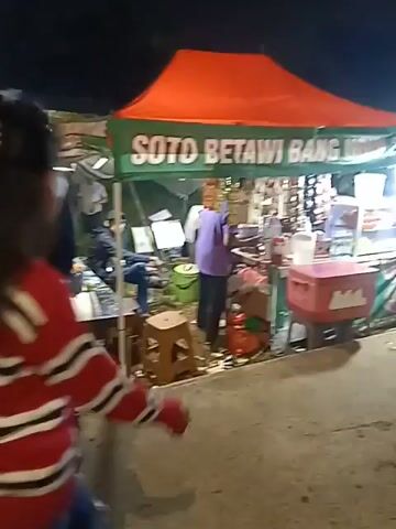 A woman wearing a red and white striped sweater and blue jeans walks away from the camera on a paved surface at night. She is walking towards a street food stall with a red canopy and green banner. There are other people around the stall and in the background, suggesting a busy night market or outdoor gathering. The atmosphere appears lively and informal.