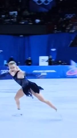 A female figure skater wearing a dark blue, sparkling, high-cut costume is performing a spin on an ice rink. She is positioned in a modified Biellmann spin, with her leg extended backward at a high angle. The skater has blonde hair and is wearing white ice skates. The background shows a blue barrier with the Olympic rings and the words "OLYMPIC WINTER GAMES."  A crowd of spectators is visible in the blurred background behind the barrier. The ice rink surface is clean and white. The camera is stationary, capturing the skater's rotation from a medium distance.