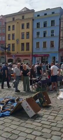 The video shows a lively outdoor market scene. A cobblestone ground indicates an old town square or historic district. Several people are browsing items displayed on tables and on the ground. In the foreground, there are cardboard boxes, one open and propped up, and a blue tarp laid out with various objects. Behind this, a group of people are gathered around tables where goods are displayed, including what appear to be glassware and other assorted items. The background features colorful historic buildings with multiple stories and windows. People are casually dressed, suggesting a daytime event. There is a sense of activity and commerce as individuals examine merchandise.