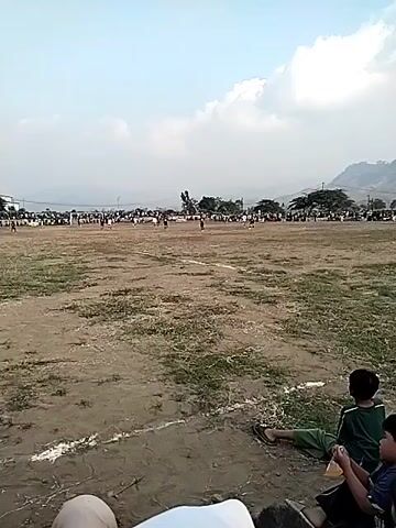 The video shows a soccer game being played on a dry, dirt field. There are spectators in the background, some sitting on benches and others standing. The sky is overcast. Players in athletic uniforms are visible on the field. The camera is positioned low, near the ground, suggesting it is being held by someone sitting or kneeling. Two young boys are visible in the foreground, with one wearing a green shirt and the other a dark shirt. One boy is holding a light-colored object. The game appears to be in progress, with players moving around the field.