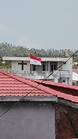 A red and white Indonesian flag flies proudly on a building with a white facade and a cross symbol. The building appears to be a church or a religious institution, with a balcony overlooking a lush, green landscape. The foreground shows a series of red-tiled roofs of residential houses, with power lines crisscrossing the frame. The sky is a pale blue, with a few wispy clouds. The overall scene suggests a peaceful, possibly rural or suburban setting in Indonesia.