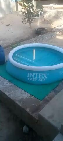 The video shows an inflatable swimming pool. The pool is light blue with a white inflatable ring around the top. The brand "INTEX" and the text "EASY SET" are visible on the side of the pool. The pool is filled with water, which appears clear. A green tarp is placed underneath the pool. The pool is situated outdoors on a dirt or gravel surface, with some concrete structures in the foreground. There is a large blue barrel to the left of the pool and some vegetation and a tire in the background. The scene appears to be a backyard or outdoor recreational area.