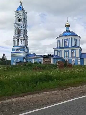 This video shows a blue and white church with a tall bell tower. The church has a blue domed roof and the bell tower is also topped with a blue dome. In front of the church is a field of green grass and wildflowers, with some piles of bricks visible in the foreground. The sky is cloudy. The camera appears to be moving, as evidenced by the slight blur in the foreground.