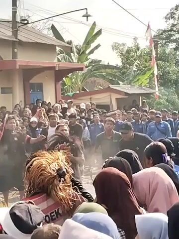 A crowd of people are gathered outdoors, watching a performance. In the foreground, a person wearing a lion-like costume with a long, furry mane and a red and white striped garment is the center of attention. Smoke or dust is visible around the performer. Several people are interacting with or observing the performer closely, some appear to be helping or guiding them. Further back, a larger crowd of spectators stands observing the event. The environment appears to be a village or residential area, with buildings and trees visible in the background. An Indonesian flag is also visible. The atmosphere suggests a cultural or celebratory event.